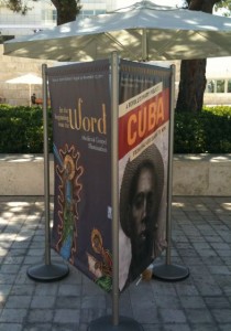 The image shows a three-sided display stand located outdoors on a paved area with trees and greenery in the background. The display features two posters. The poster on the left side has the text "The Word" and appears to be related to a religious or gospel theme, with an image of a religious figure. The poster on the right side has the text "CUBA" and features a black-and-white photograph of a person wearing a hat. The display is set up under a large umbrella providing shade.