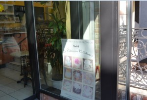 The image shows the exterior of a shop with a glass window. Inside the window, there is a sign with images of various cupcakes or pastries and text in French. The interior of the shop is visible through the window, showing a plant and some furniture. The reflection on the glass also shows part of the street or building outside.