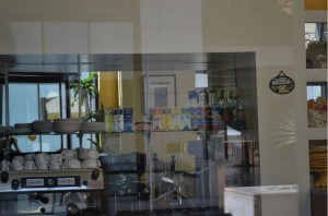 The image shows the interior of a kitchen or café, viewed through a glass window. There are shelves with various items, including boxes and bottles, and a coffee machine with cups stacked on top. The reflection on the glass shows parts of the room and possibly some outdoor scenery. The overall setting appears to be clean and organized.