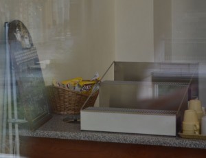 The image shows a countertop with a few items on it. There is an empty display case, a basket containing some packaged snacks, and a few disposable cups stacked on the right side. To the left, there is a menu board or sign. The scene appears to be inside a small shop or café.