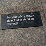 A rectangular sign with white text on a dark background is placed on a stone surface. The sign reads, "For your safety, please do not sit or stand on the wall."