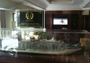 The image shows a detailed model of a cruise ship encased in a glass display. Above the display case, there is a sign with the text "Utopia" and "Residential Holiday Liner Visiting World Famous Events." In the background, there is a television mounted on the wall, displaying an image of a person on a beach. The room appears to be well-lit with natural light coming through the windows.