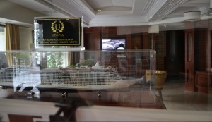 The image shows a detailed model of a cruise ship displayed inside a glass case. The model is situated in a well-lit room with large windows and a television screen in the background. Above the model, there is a sign with the word "UTOPIA" and some additional text. The room has a sophisticated and elegant interior design with wooden elements and comfortable seating.