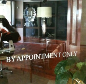 The image shows the interior of an office or a professional space viewed through a glass door. The glass door has the text "BY APPOINTMENT ONLY" written on it. Inside, there is a wooden desk, a chair, a lamp, and some plants. The word "UTOPIA" and a logo with the letter "U" surrounded by a laurel wreath are also visible on the glass door.