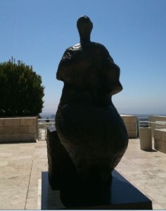 A large, abstract bronze sculpture is displayed outdoors on a stone platform. The sculpture appears to be a stylized human figure with a rounded, voluminous form. In the background, there is a clear blue sky, a tree, and some architectural elements, including a railing and stone walls. The setting suggests an open, public space, possibly a museum or a park.