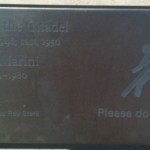 The image shows a dark-colored plaque with engraved text. The text includes names and dates, and there is a small figure of a person walking with a cane on the right side. The text also includes a message that says "Please do" but the rest of the message is not fully visible.