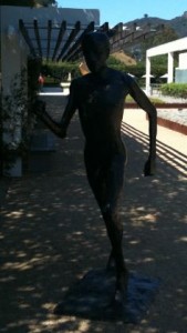 The image shows a dark bronze statue of a person in a running pose. The statue is located outdoors, under a pergola that casts shadows on the ground. In the background, there are some buildings and greenery, with a hill visible in the distance. The overall setting appears to be a garden or a courtyard.