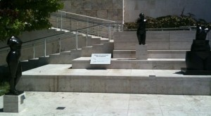 The image shows an outdoor area with three black sculptures of human figures placed on different levels of a stone staircase. The sculptures appear to be abstract representations of human forms. There is a sign placed on one of the steps, but the text on the sign is not clearly readable. The area is surrounded by a stone wall and there is a metal handrail along the staircase. Some greenery is visible in the background.