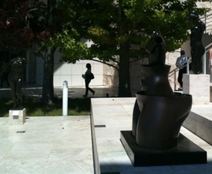 The image shows an outdoor area with several abstract sculptures. In the foreground, there is a large, dark-colored abstract sculpture on a pedestal. In the background, there are more sculptures and a few people walking or standing near the sculptures. The area is surrounded by trees and a building with a light-colored facade. The overall scene appears to be a sculpture garden or an outdoor art exhibit.