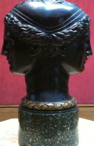 A sculpture of a two-faced head, likely representing the Roman god Janus, is shown. The sculpture is dark in color and features detailed hair and facial features on both faces, which are positioned back-to-back. The base of the sculpture is a dark, speckled pedestal. The background is a solid red color.