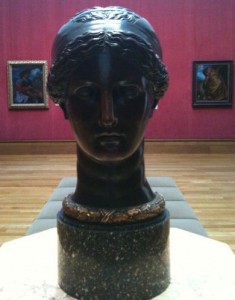 A dark bronze bust of a woman's head is displayed on a marble pedestal in an art gallery. The gallery has wooden floors and red walls, with framed paintings hanging in the background. The bust features detailed hair and a serene facial expression.