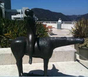 A bronze statue of a child riding a horse is displayed outdoors on a sunny day. The child is sitting upright on the horse, which has an elongated neck and a stylized, abstract form. The background features a terrace with plants, a bench, and a person standing near a railing, with mountains visible in the distance.