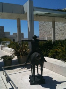 The image shows a bronze statue of a person riding a horse. The statue is placed in an outdoor area with modern architectural elements, including white stone walls and columns. There are potted plants and greenery around the statue, and the sky is clear and blue.