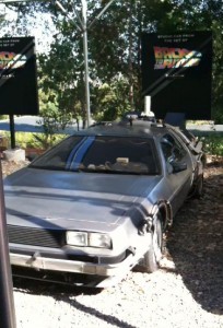 A silver DeLorean car is parked outdoors on a gravel surface. The car has various modifications and equipment attached to it, resembling the time machine from the "Back to the Future" movie series. In the background, there are two signs with the "Back to the Future" logo. Trees and greenery are visible behind the signs.