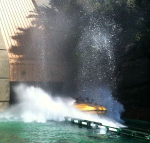 A yellow boat is making a big splash as it descends a water ride, creating a large spray of water. The boat is on a track and surrounded by green water, with a tunnel entrance and trees in the background.