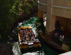 The image shows a water ride at an amusement park. A boat filled with people is moving along a narrow waterway surrounded by lush greenery. The boat is yellow with black seats, and the passengers appear to be enjoying the ride. To the right, there is a building structure with a few people standing on a balcony, possibly waiting for their turn or watching the ride. The water in the canal is green, reflecting the surrounding vegetation.