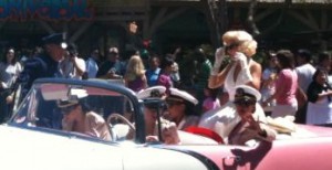 A group of people are participating in a parade. They are riding in a classic pink and white convertible car. Some of the individuals are dressed in vintage military uniforms, while a woman standing in the car is dressed in a white dress and has blonde hair styled in a retro fashion. There are spectators in the background watching the parade.