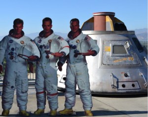 Three astronauts in spacesuits are standing in front of a space capsule. The astronauts are wearing white suits with various patches and insignias, and they are holding helmets. The space capsule behind them is metallic and has a conical shape with a small window and various markings. The background shows a clear sky and some distant mountains.