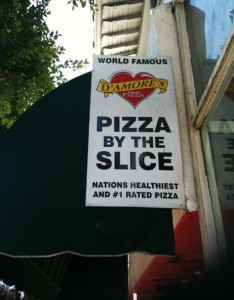 A sign for D'Amore's Pizza is displayed. The sign reads "WORLD FAMOUS D'AMORE'S PIZZA" at the top, followed by "PIZZA BY THE SLICE" in large bold letters. Below that, it states "NATION'S HEALTHIEST AND #1 RATED PIZZA." The sign is attached to a building with a green awning and is partially shaded by nearby trees.