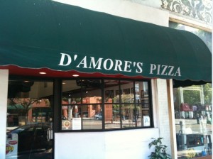 The image shows the exterior of a pizza restaurant with a green awning that has the text "D'AMORE'S PIZZA" written on it in white letters. The restaurant has large windows, and some reflections of the street and buildings can be seen in the glass. There is also a small plant near the entrance.