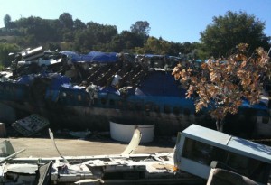The image shows the wreckage of a large airplane, which appears to have crashed. The fuselage is heavily damaged and broken into pieces, with debris scattered around the site. There are trees and some vegetation in the background, indicating that the crash site is in an outdoor area. The sky is clear and blue, suggesting it is a sunny day.