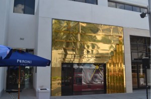 The image shows the exterior of a building with a large, reflective gold facade. There is a storefront with a window display partially covered by a white sheet. To the left of the building, there is a blue umbrella with the word "PERONI" written on it, indicating a nearby outdoor seating area. The building has a modern design with clean lines and large windows. The address number "342" is visible on the left side of the building.