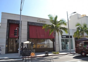 The image shows a street view of a Cartier store with large red awnings and a palm tree in front. Next to the Cartier store is another store with a sign that reads "ELOGE." There is a bus or large vehicle parked on the street to the right. The scene is set on a sunny day with clear skies.