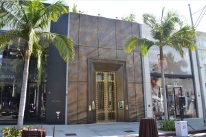 The image shows the exterior of a high-end retail store with a large, ornate golden door in the center. The building has a modern design with a sleek, dark facade. There are two palm trees in front of the store, and the sidewalk is clean and well-maintained. The store to the right has large glass windows displaying mannequins dressed in fashionable clothing.