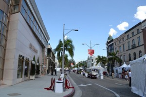 A street scene with a clear blue sky. The street is lined with palm trees and buildings on both sides. On the left, there is a large building with a sign that reads "BVLGARI." On the right, there are several white tents set up along the sidewalk. There are a few people walking and some cars parked along the street. The overall atmosphere appears to be calm and sunny.