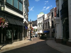 A narrow, cobblestone street lined with elegant buildings featuring large windows and awnings. The buildings have a mix of architectural styles, including arched windows and decorative columns. Hanging flower baskets adorn the street, and there are a few street lamps. The sky is clear and blue, suggesting a sunny day. The street appears to be part of a shopping or commercial area.