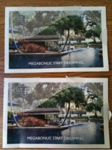 The image shows two identical postcards placed one above the other on a wooden surface. Each postcard features a serene poolside scene with palm trees, a modern building, and lush greenery in the background. The text "MEGABONUS: START DREAMING." is printed at the bottom of each postcard.