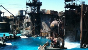 The image shows a water-based stage set, likely for a live-action show or movie scene. The set includes various industrial structures, such as towers, platforms, and machinery, all with a weathered, post-apocalyptic appearance. There are boats and people in the water, suggesting an action sequence or performance. The water is a bright blue, contrasting with the dark, rusty structures.