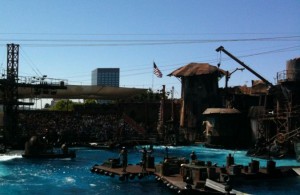 The image shows a water-based stage set, likely for a live-action show or performance. The set includes various structures such as platforms, towers, and cranes, with an American flag visible in the background. There is a large pool of water in the foreground, and a crowd of spectators can be seen seated in the background, watching the performance. The sky is clear and blue, indicating a sunny day.