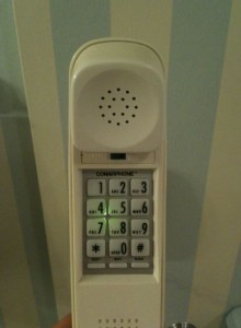 The image shows a close-up of a white cordless telephone with a numeric keypad. The phone is vertically oriented, and the earpiece with speaker holes is at the top. The keypad includes numbers 0-9, along with the star (*) and pound (#) symbols. The number 4 key is illuminated with a green light. The background features a wall with vertical stripes in light blue and white.