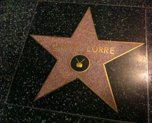 The image shows a star on the Hollywood Walk of Fame. The star is pink with a gold border and is embedded in a black terrazzo floor. It has the name "CHUCK LORRE" inscribed on it, along with a symbol of a television, indicating recognition in the television industry.