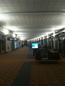 The image shows an empty airport terminal with rows of seats and illuminated signs. The terminal has a high ceiling with bright lights, and there are digital screens and advertisements along the walls. The floor is carpeted with a patterned design, and the overall atmosphere appears quiet and calm.