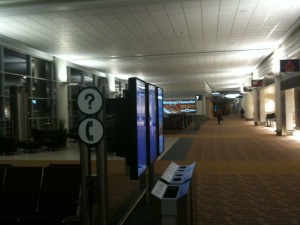 The image shows an empty airport terminal with a long corridor. There are information signs with a question mark and a telephone symbol on the left side. The terminal is well-lit with overhead lights, and there are seating areas along the sides. In the distance, there are more signs and a few people walking. The overall atmosphere is quiet and calm.