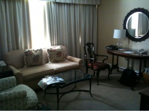 The image shows a cozy living room with a beige sofa adorned with two floral-patterned cushions. In front of the sofa is a glass coffee table with a magazine on it. To the left of the sofa is an armchair with a patterned fabric. On the right side of the room, there is a wooden desk with a chair, a table lamp, and some items on the desk, including a laptop. Above the desk is a round mirror with a decorative frame. The room has beige walls and is lit by natural light coming through a window with closed curtains. There is also a black backpack on the floor near the desk.