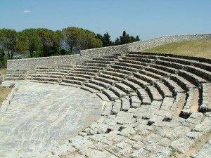 The image shows an ancient stone amphitheater with tiered seating. The structure is made of stone and appears to be weathered, indicating its age. In the background, there are trees and a clear blue sky. The amphitheater is situated outdoors, and the seating is arranged in a semi-circular pattern.