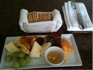 A rectangular white plate with a variety of cheeses, green and red grapes, dried apricots, and a small bowl of honey. Behind the plate, there is a small white basket filled with crackers. To the right, there are two folded white napkins with a fork and knife placed on top. The setting is on a dark countertop.