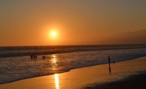 The image shows a serene beach scene at sunset. The sun is low on the horizon, casting a warm, golden glow over the water and the sandy shore. There are a few people in the water and one person standing on the wet sand, enjoying the tranquil moment. The sky is clear with a gradient of colors from orange to blue as the day transitions into evening.