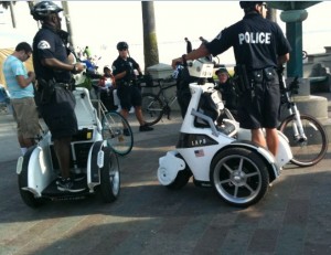 The image shows several police officers patrolling an outdoor area. Two of the officers are riding Segways, while others are on bicycles. The officers are wearing uniforms with the word "POLICE" visible on their backs. There are also a few civilians in the background, one of whom appears to be using a smartphone. The scene takes place in a public space with some trees and benches visible.