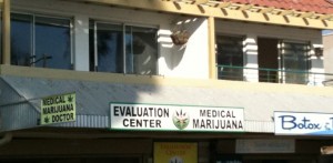 The image shows the exterior of a building with signs indicating it is a medical marijuana evaluation center. The signs read "MEDICAL MARIJUANA DOCTOR," "EVALUATION CENTER," and "MEDICAL MARIJUANA." There is also a partial sign on the right that reads "Botox." The building has large windows and a balcony on the upper floor.