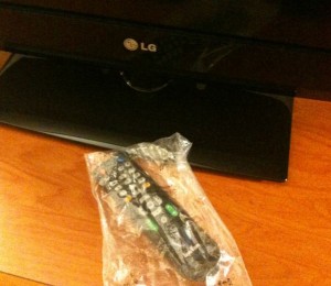 The image shows an LG television placed on a wooden surface. In front of the television, there is a remote control wrapped in a plastic bag.