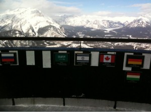 The image shows a scenic view of snow-covered mountains in the background. In the foreground, there is a railing with several plaques displaying different national flags, including those of Russia, Saudi Arabia, Israel, Canada, Germany, and Hungary. The plaques likely indicate the direction and distance to these countries from the viewpoint.