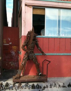 A large, rusted metal sculpture of a humanoid figure stands against a red wooden building with a window. The sculpture has an elongated head and appears to be made from various metal parts. In front of the sculpture, there are several smaller metal figurines arranged on the ground. The scene is outdoors, and the building has a partially open window with a reflection of the sky.