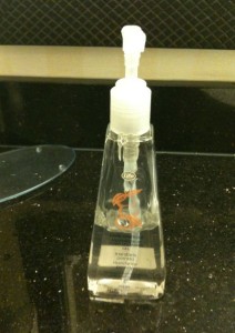 The image shows a clear plastic pump bottle filled with hand soap or sanitizer. The bottle is placed on a black countertop, and there is a small label on the front of the bottle. The background includes a tiled wall and part of a glass shelf.