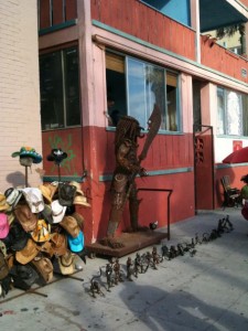 The image shows a street scene with a large metal sculpture of a humanoid figure holding a large blade, positioned in front of a building with red and pink walls. To the left of the sculpture, there is a display of various hats hanging on a stand. On the ground in front of the sculpture, there are several smaller metal sculptures arranged in a line. The building has large windows and appears to be a shop or a similar establishment.