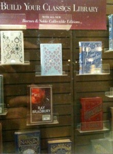 The image shows a display case with a sign at the top that reads "Build Your Classics Library with all-new Barnes & Noble Collectible Editions." The display contains several classic books, including:

- A book with a white cover and intricate blue designs.
- A book with a light blue cover and a pattern of small designs.
- A book titled "Pride and Prejudice" with a blue cover.
- A book by Ray Bradbury with a red and black cover.
- A book with a red cover and gold designs.
- A book from "The Chronicles of Narnia" series with a blue cover.

The books are arranged on clear shelves within the display case.