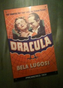 The image is a vintage movie poster for the film "Dracula" featuring Bela Lugosi. The poster has an orange and purple color scheme with a web-like background. At the top, it reads "The Vampire Bat That Lives on Human Blood!" Below this, there is an image of two characters, one appearing to be Dracula and the other a woman. The title "DRACULA" is prominently displayed in large letters, with "BELA LUGOSI" written underneath. At the bottom, it states "PREMIERED 1931."